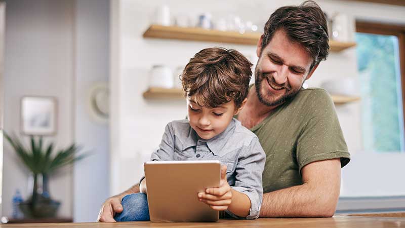 Pai e filho pequeno, apoiados numa mesa, com um tablet na mão